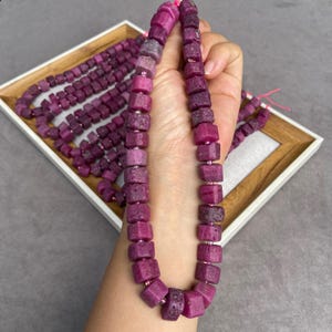 Peut inclure: Un collier de perles de rubis brutes en forme de cube, d'une teinte magenta-rose profonde. Les perles présentent une surface naturelle texturée et sont enfilées sur un fin cordon rose, formant une ligne continue de segments de pierre.
