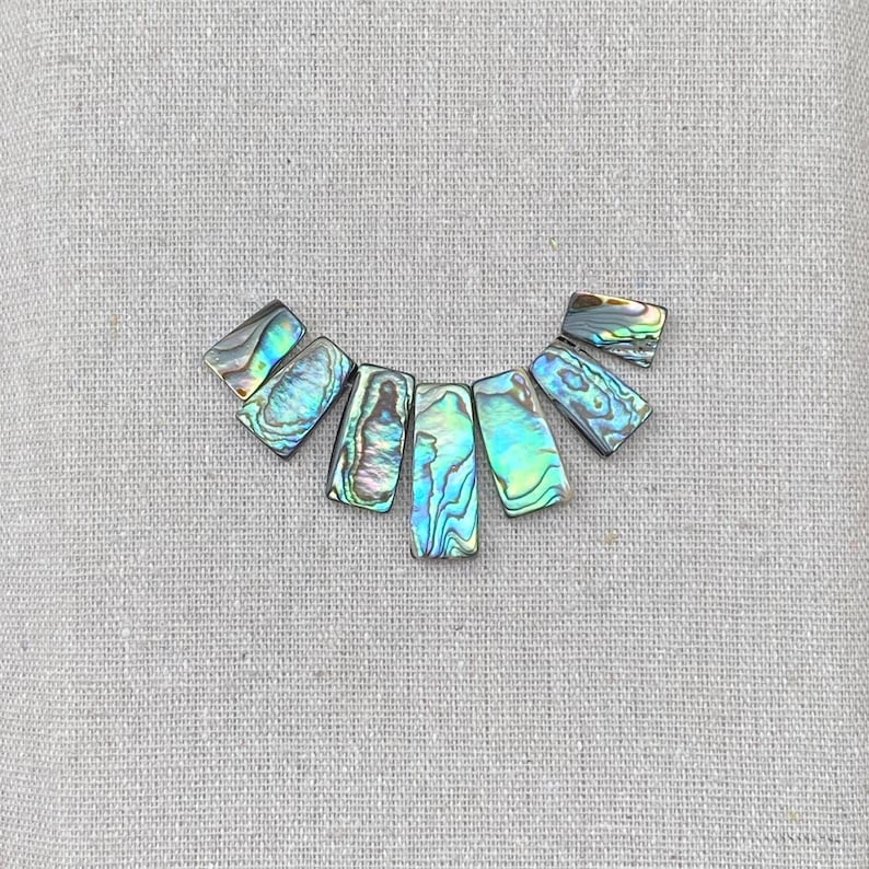 以下が含まれることがあります： 半円状に配置された7つの長方形のアワビの貝殻のピース。各ピースは、青、緑、紫の色合いの渦巻き模様を持つ磨かれた虹色の表面が特徴で、貝殻素材の自然な光沢を示しています。