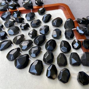 May include: Strands of faceted black gemstone beads. The beads have irregular shapes and are strung on a clear cord. The beads are displayed on a white surface inside a wooden tray.