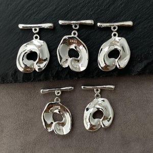 Può includere: Cinque chiusure a T in argento per la creazione di gioielli, ognuna con un pendente circolare irregolare dallo stile organico e fuso, fissato a una barra orizzontale. Ogni pezzo reca il marchio S925, che indica la composizione in argento sterling.