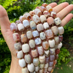 Op de afbeelding: Een meerstrengs ketting met tonvormige kralen in crème, perzik en bruin. De kralen zijn geregen met goudkleurige metalen accenten, wat een natuurlijke uitstraling creëert. De ketting wordt in een hand gehouden.
