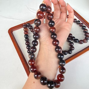 Puede incluir: Un conjunto de collar y pulsera con grandes cuentas redondas pulidas en tonos rojo oscuro y marrón. Las cuentas están ensartadas, creando un contraste llamativo. Las joyas se exhiben en una bandeja de madera.