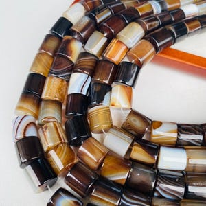 Può includere: Primo piano di una collana di perline di agata. Le perline sono cilindriche con un mix di colori marrone, beige e bianco. Le perline hanno una superficie lucida e liscia e sono infilate, pronte per la creazione di gioielli.
