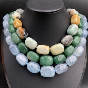 Può includere: Collana multistrato con grandi pietre preziose lucide nei toni del blu, verde, giallo e marrone. Le pietre sono infilate su un cordino blu, creando un effetto a strati. La collana è esposta su un busto di gioielleria nero.