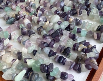 May include: A collection of tumbled fluorite gemstones in shades of purple, green, and white. The stones are irregular in shape and size, with a polished, glossy finish. The gemstones are arranged closely together, creating a textured surface.