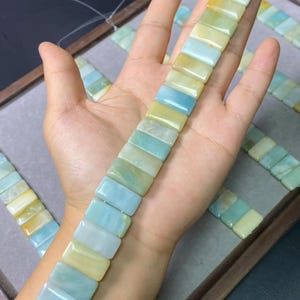 May include: A string of rectangular gemstone beads in pale yellow, blue, and green hues. The beads are held in a hand, with more strands visible in the background. The beads have a polished, smooth surface.