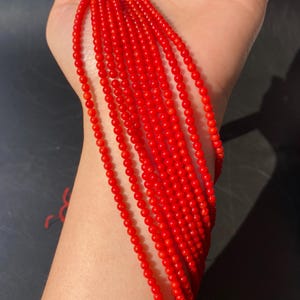 May include: A collection of vibrant red coral beads, strung together to form multiple strands. The beads are small and round, with a consistent size and color. The strands are held in a person's hand, showcasing the beads' rich color and texture. The beads are likely for jewelry making.
