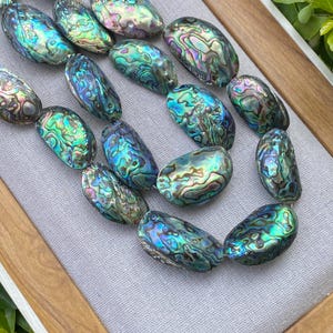 May include: A close-up of a necklace made of iridescent abalone shell beads. The beads are oval-shaped with a mix of blue, green, and purple hues. The necklace is displayed on a gray fabric inside a wooden tray.