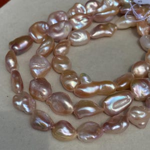 Könnte beinhalten: Eine Kette aus unregelmäßig geformten, irisierenden Süßwasserperlen. Die Perlen haben eine zarte rosa- und pfirsichfarbene Farbe mit einem glänzenden Glanz. Die Perlen sind aufgefädelt und bilden eine Halskette oder ein Armband.