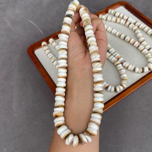 Puede incluir: Un largo collar de cuentas circulares de bordes planos hechas de material de concha natural. Las cuentas presentan un acabado liso y pulido con una paleta de colores que va desde el blanco crema hasta tonos bronceados y marrones, ensartadas en un hilo blanco.