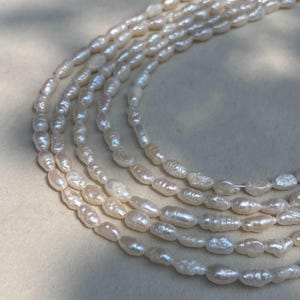 Puede incluir: Un collar de múltiples hilos compuesto por numerosas perlas de agua dulce pequeñas, irregulares y de color blanco hueso. Las perlas presentan un acabado lustroso e iridiscente con formas y tamaños orgánicos variados, ensartadas en varios bucles.