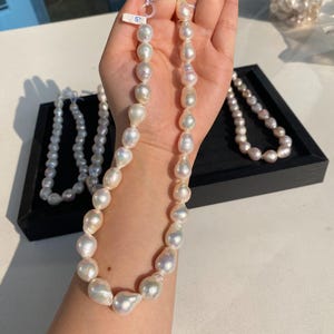 Puede incluir: Una colección de collares de perlas blancas. Un collar se sostiene en una mano, mostrando perlas grandes e irregulares. Otros collares se exhiben en una bandeja negra. Las perlas tienen un brillo lustroso, reflejando la luz.