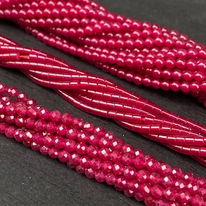 May include: Close-up of several strands of faceted, deep red beads. The beads vary in shape, including small, round, and faceted, with a glossy finish. The strands are arranged in a slightly overlapping pattern, showcasing the color and texture.