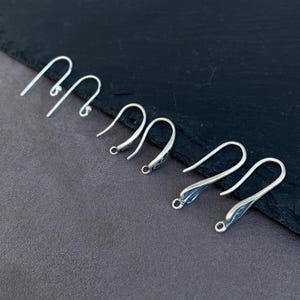 Puede incluir: Tres pares de ganchos para pendientes de metal plateado, cada uno con un diseño curvo y alargado con una pequeña anilla en la parte inferior para sujetar componentes de joyería. Los ganchos tienen un acabado liso, pulido y funcional.