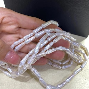 Puede incluir: Un collar compuesto por numerosas perlas blancas y oblongas. Las perlas tienen un brillo lustroso y están ensartadas para formar un hilo continuo. El collar se muestra sobre un fondo neutro.