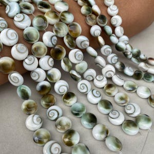 Könnte beinhalten: Nahaufnahme von mehreren Strängen Perlen. Die Perlen sind eine Mischung aus Weiß, Olivgrün und Braun, mit einem Spiralmuster. Sie sind aufgefädelt, wahrscheinlich für Schmuck oder Bastelarbeiten. Die Perlen haben eine polierte, glänzende Oberfläche.