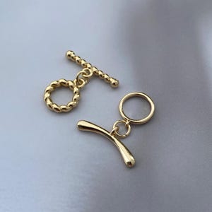 May include: Two gold-toned toggle clasps for jewellery making. One clasp features a twisted rope texture on both the circular ring and the bar component. The second clasp consists of a smooth, polished circular ring and a curved, solid bar component.