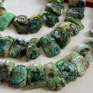 Op de afbeelding: Een ketting bestaande uit ruwe, gefacetteerde groene edelsteenkralen met gevarieerde texturen en natuurlijke insluitingen. De stenen vertonen tinten bosgroen, olijf en mos, geregen aan een dun groen koord voor een robuuste uitstraling.