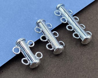 May include: Three silver-toned cylindrical jewelry clasps arranged diagonally. The smallest has two loops, the middle has three loops, and the largest has four loops on each side. Each clasp features a smooth, polished metallic finish.