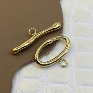 Può includere: Un set di chiusura a T color oro composto da una barra allungata e liscia con un occhiello centrale e un anello ovale abbinato con un piccolo occhiello attaccato. Entrambi i componenti presentano una finitura metallica lucida e riflettente.