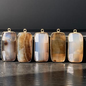 May include: Five rectangular gemstone pendants with gold-coloured metal frames. The stones vary in colour, including shades of brown, grey, and white. Each pendant has a small loop at the top for hanging.