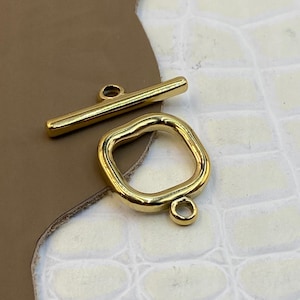 Pode incluir: Um fecho de metal dourado para bijuteria, composto por uma barra alongada e lisa com um laço central e um anel quadrado irregular separado, apresentando um pequeno laço de fixação na borda inferior.