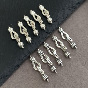 Può includere: Una collezione di nove chiusure per gioielli in metallo color argento, ciascuna dotata di un meccanismo a gancio con un'estremità cilindrica scanalata e un marchio S925 stampato sulla maglia centrale, disposte in modo sparso.