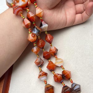 Peut inclure: Gros plan sur des perles de pierres précieuses orange, marron et blanches. Les perles à facettes sont enfilées sur un cordon rouge et vert. Les pierres précieuses ont une forme géométrique et sont disposées en collier ou bracelet.