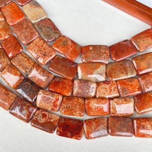 Puede incluir: Primer plano de un collar de cuentas rectangulares de coral. Las cuentas son una mezcla de tonos rojo-naranja y marrón, con patrones visibles. Las cuentas están ensartadas, lo que sugiere que son para hacer joyas o un collar terminado.