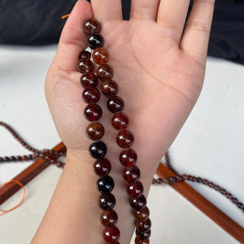 May include: Strands of polished, round gemstone beads in shades of deep red, brown, and black. The beads are held in a hand, showcasing their smooth, reflective surfaces. The beads are strung together, suggesting jewelry or craft supplies.