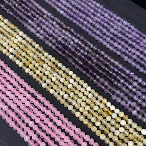 May include: Close-up of several strands of gemstone beads in various colors. The beads are arranged in parallel rows, showcasing shades of pink, yellow, and purple. The beads appear to be small and faceted, reflecting light. The background is a dark, solid color.
