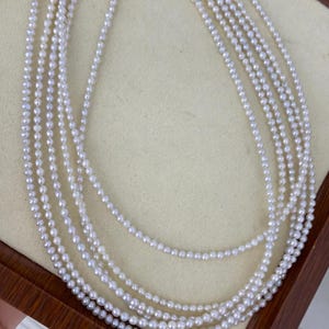 Könnte beinhalten: Eine mehrsträngige Perlenkette. Die Halskette besteht aus mehreren Strängen kleiner, runder, weißer Perlen. Die Stränge sind geschichtet und erzeugen einen Kaskadeneffekt. Die Perlen haben einen subtilen Glanz, und die Halskette wird auf einer hellen Oberfläche präsentiert.