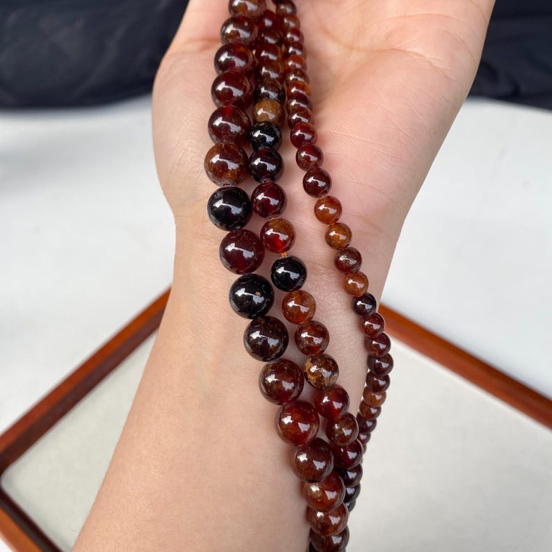 May include: Three strands of gemstone beads in varying sizes and shades of red and brown. The beads are round and appear to be polished. The strands are held in a hand, with a wooden frame in the background.