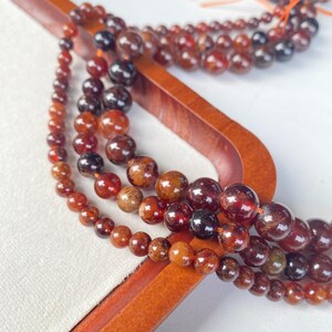 May include: Several strands of gemstone beads in varying sizes and shades of red, brown, and black are displayed. The beads are strung on orange thread and arranged on a wooden display piece. The beads have a polished, glossy finish.