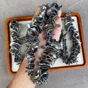 Könnte beinhalten: Ein Strang aus dunklen, irisierenden Muschelperlen mit unregelmäßigen, gerüschten und blütenblattartigen Formen. Die Perlen weisen einen metallischen Glanz mit tiefgrauen, anthrazitfarbenen und bronzenen Tönen auf und sind auf einer dünnen Schnur.