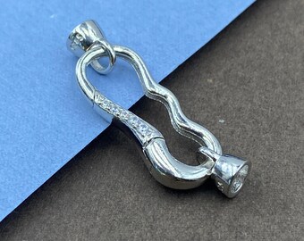 May include: A silver-toned metal jewelry clasp featuring a wavy, elongated oval shape. The clasp is accented with a row of small, clear, sparkling stones along one side and includes a small loop at the top for attachment to a necklace or bracelet.