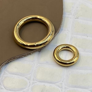 May include: Two polished gold-tone metal rings with a smooth, rounded surface. The larger ring is positioned to the left, while the smaller ring is placed to the right. Both rings feature a seamless, circular design with a reflective finish.
