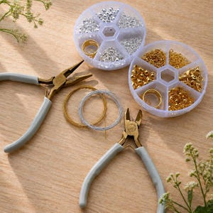 May include: A selection of jewellery-making supplies, including two pairs of grey-handled pliers, two clear plastic containers filled with silver and gold beads, and spools of gold and silver wire. The items are arranged on a light brown wooden surface.
