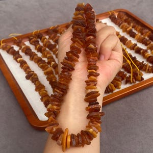 Può includere: Una collana di perle d'ambra grezze e irregolari in tonalità giallo oro, arancione e marrone scuro. I frammenti di pietra traslucida e levigata sono infilati su un sottile cordino giallo, mostrando variazioni naturali di forma e colore.