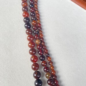 May include: Close-up of several strands of gemstone beads in varying sizes and shades of red, brown, and black. The beads have a polished, glossy finish and are strung together, creating a layered effect. The background is a soft, neutral color.