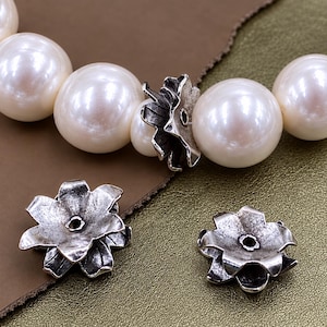 Può includere: Una collana di grandi perle rotonde color bianco sporco alternate a copriperle in argento ossidato a forma di fiore. Due ulteriori copriperle in argento ossidato a forma di fiore sono posizionati separatamente, ciascuno con un foro centrale.