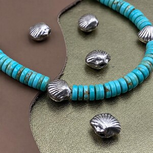 Op de afbeelding: Een ketting bestaande uit een streng kleine, ronde, turquoise schijfkralen met bruine matrixpatronen, afgewisseld met metallic zilverkleurige schelpvormige kralen. Verschillende losse zilveren schelpvormige kralen liggen verspreid.