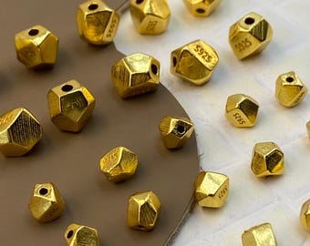 May include: A collection of small, gold-colored, multi-faceted beads with a textured surface. Each bead features a central hole and is stamped with the text S925, indicating the metal composition. The beads have an irregular, geometric shape.