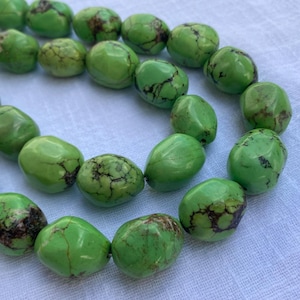 Puede incluir: Una colección de cuentas de color verde lima, lisas y redondeadas, con vetas de color marrón oscuro. Cada cuenta presenta un acabado pulido y formas orgánicas irregulares, dispuestas en un grupo suelto para mostrar el color vibrante.