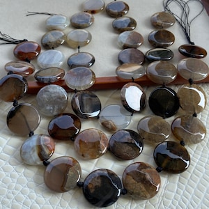 Può includere: Una collezione di perline di agata circolari levigate in tonalità marrone, marrone chiaro, grigio e bianco traslucido. Le pietre lisce, piatte e a forma di moneta sono infilate su filo nero, mostrando venature naturali e vari motivi cromatici.