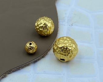 May include: Three spherical beads with a gold-toned finish and a hammered, textured surface. The beads vary in size, with one small bead and two larger beads, each featuring a central hole for threading through a cord or wire.