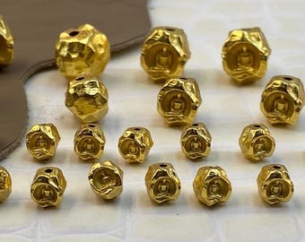 May include: A collection of gold-colored beads with irregular, textured surfaces. Several beads feature a recessed, centered carving of a seated figure in a meditative pose. Each bead has a small hole at the top for stringing purposes.