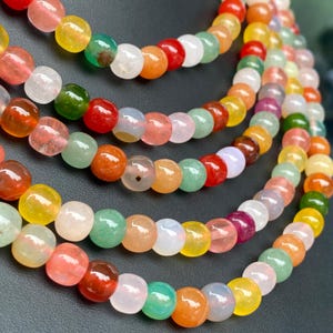 Può includere: Una collana a più fili composta da perline lisce, rotonde e traslucide in una vivace gamma di colori tra cui rosso, arancione, giallo, verde e bianco. Le perline sono infilate in diverse file a cascata, creando un aspetto stratificato.