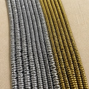 Puede incluir: Filas de cuentas de color plata y oro. Las cuentas tienen una superficie acanalada y texturizada y están dispuestas en líneas paralelas. El fondo es de color beige neutro. Las cuentas podrían utilizarse para la fabricación de joyas u otros proyectos de artesanía.