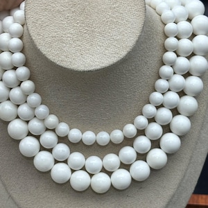 May include: A multi-strand white beaded necklace displayed on a mannequin. The necklace features graduated sizes of round beads, creating a layered effect. The beads have a glossy finish and are arranged in a cascading design.